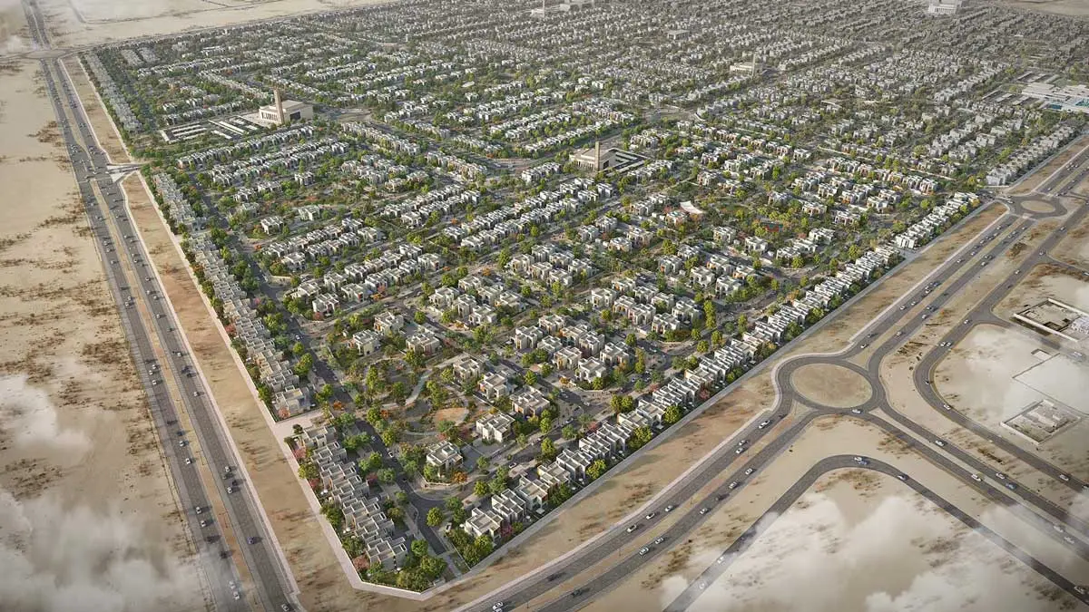 Aerial view of a large low-rise residential community with rows of villas and townhouses, tree-lined streets, mosques, and a road roundabout at the community entrance, set against surrounding desert landscape.