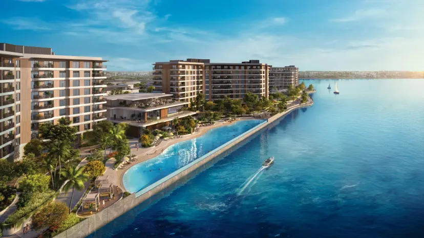 Aerial view of Gardenia Bay residential towers along the waterfront, with an infinity pool and beach club edging the water, palm-lined promenade, and boats passing on the sea.